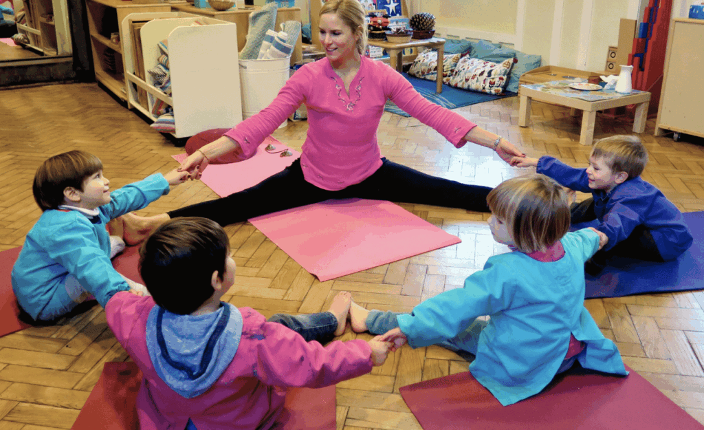 Children sit in a circle holding hands with their instructor, participating in a yoga-inspired movement session on colourful mats indoors.